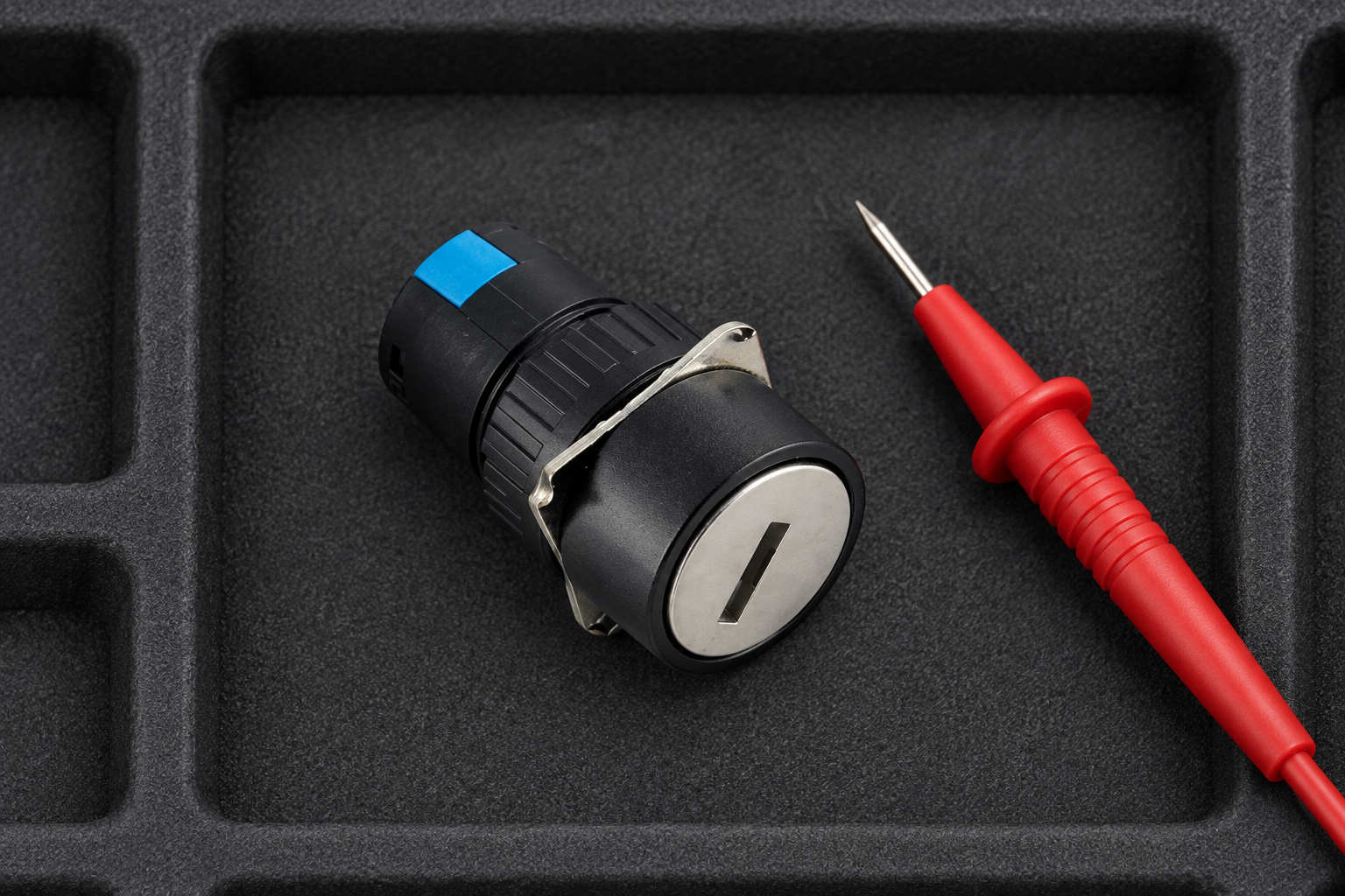 12V and 24V Key Switches: Contact Duty and Input Selection body2 Black keyed operator with silver keyslot face and original blue rear segments, disconnected on a padded tray beside a meter probe that touches no terminal